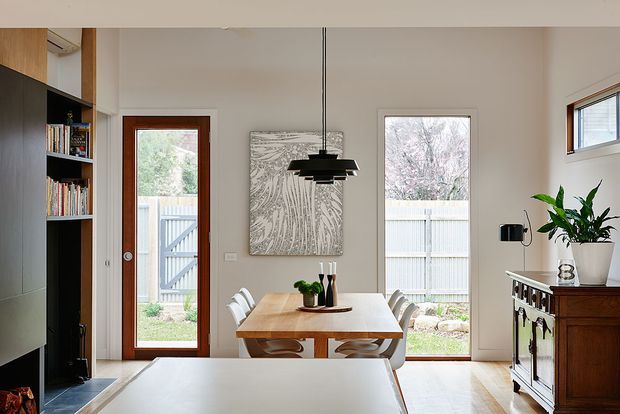 Bright contemporary dining area viewed from an adjacent kitchen or living space. A light timber dining table with curved chairs sits beneath a black tiered pendant light, with a large monochrome artwork on the wall. Tall glazed doors and windows bring in daylight and overlook a fenced garden. Built-in dark shelving and cabinetry are visible at left, while a dark timber sideboard and indoor plant sit at right.