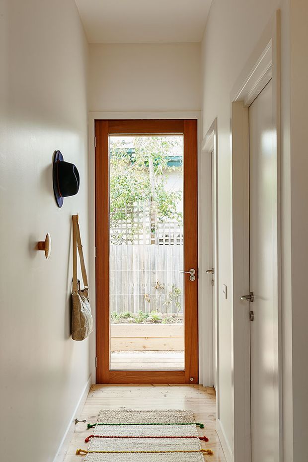 A narrow, bright residential hallway leading to a glazed timber entry door. The corridor has warm white walls and ceiling, pale timber floorboards, white interior door trim and hardware, wall-mounted hooks with a hanging bag, and a simple striped runner. Through the glass entry door, a small fenced garden courtyard is visible.