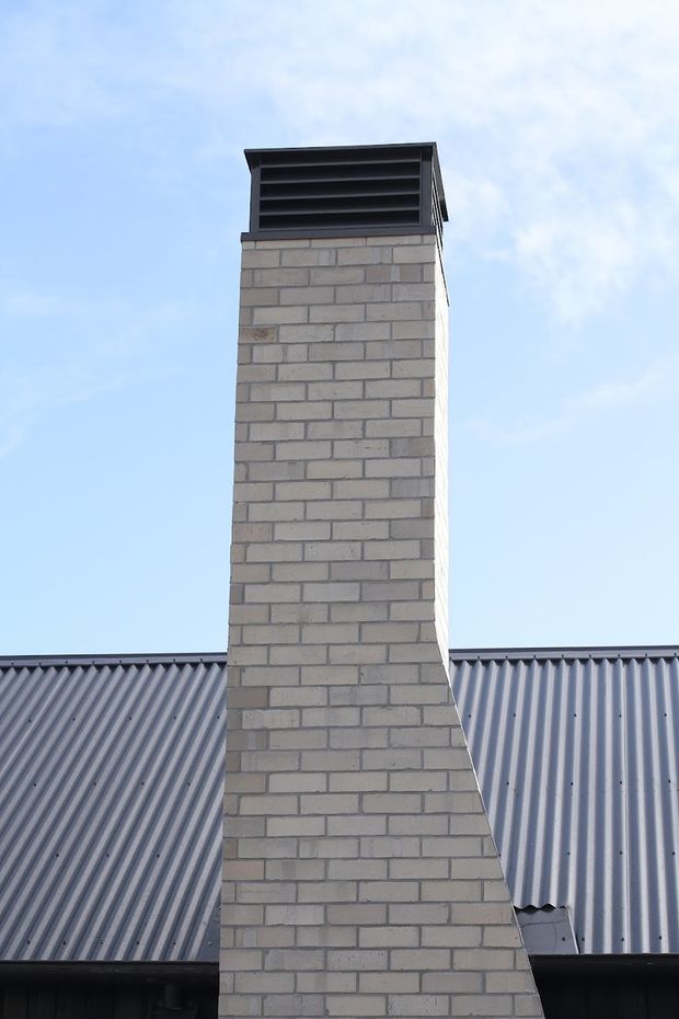 Exterior close-up of a tall light-grey brick chimney with a dark louvred metal cap, rising above a corrugated metal pitched roof. The image is dominated by the masonry chimney and roof envelope against a bright sky.