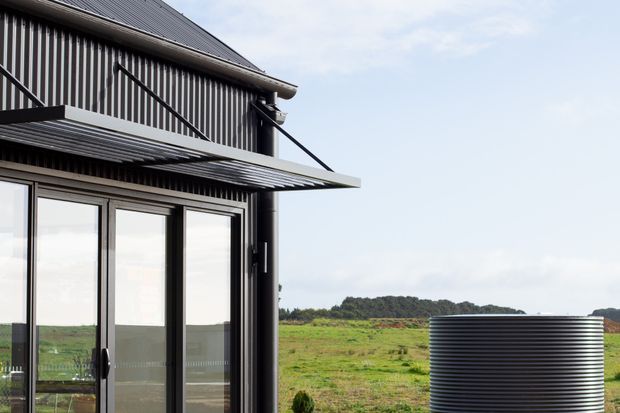 Exterior architectural detail of a dark corrugated-metal building with a pitched roof, vertical cladding, glazed sliding doors, and a projecting corrugated awning. A large ribbed cylindrical water tank sits beside the building, with open rural fields visible beyond.