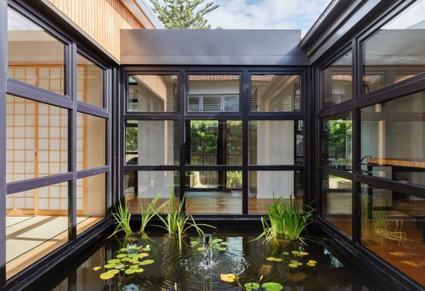 A refined enclosed courtyard or sunroom surrounding a shallow indoor water feature. Black-framed glazing forms three sides, with a timber-clad upper wall and visible Japanese-inspired screens and interiors beyond. The pond contains water lilies and grasses, creating a calm connection between the architecture and planted water feature.