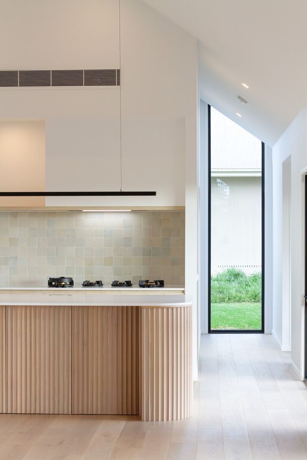A bright contemporary kitchen opening onto a light-filled hallway. The kitchen features a long gas cooktop, pale square tiled splashback, warm fluted timber cabinetry with a curved end, light timber flooring, white walls, and a slim black linear pendant. A tall glazed opening provides views to a small landscaped courtyard or garden, while the ceiling incorporates recessed lighting and linear air-conditioning grilles.