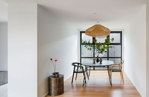 Bright contemporary dining area with a round table and mixed black and natural timber chairs, positioned beneath a large woven pendant light. The room has white walls, warm timber flooring, a black-framed glazed window with vertical screening, and simple botanical styling.