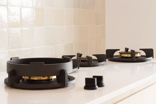 Close-up of a modern gas cooktop with three black cast-iron burner grates and brass-toned burner components, set into a white countertop. A glossy off-white square-tile splashback forms the background, with separate black control knobs visible in the foreground.