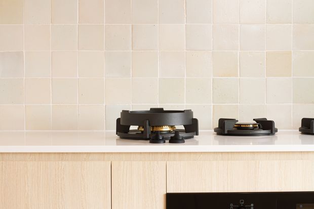 Close-up of a minimalist kitchen cooktop installation, showing black gas burners set into a white benchtop, pale cream square tiled splashback, and light timber cabinetry below. The image focuses on the cooktop and fixed kitchen finishes; only part of a black appliance is visible at the lower right.
