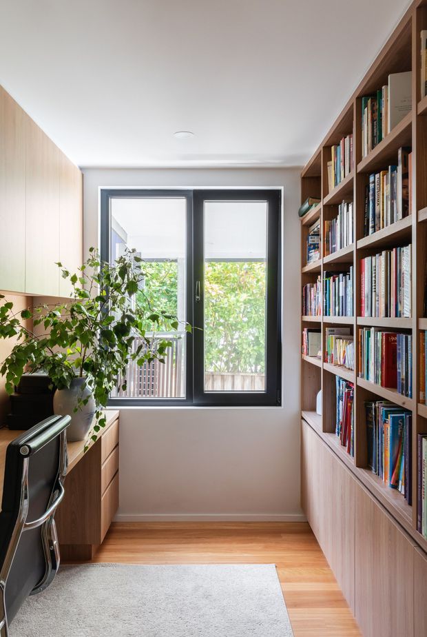 A compact, bright home office lined with extensive built-in timber cabinetry and open bookshelves. A black-framed glazed door or window opens toward leafy greenery, while a desk, office chair, potted plant, timber flooring, and a pale area rug create a warm, functional workspace.