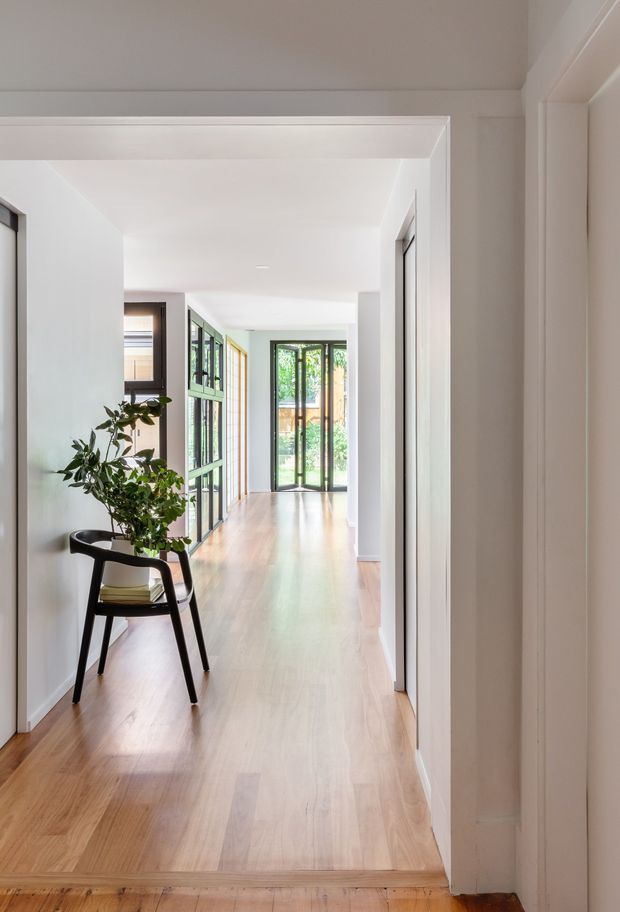 A bright, elongated residential hallway with pale timber flooring, white walls and ceiling, and multiple internal doorways. Black-framed glazed doors and windows at the far end bring in abundant daylight and greenery. A simple black chair with a potted leafy plant and books forms a small vignette along the left wall. The space has clean contemporary detailing with a restrained, calm palette.