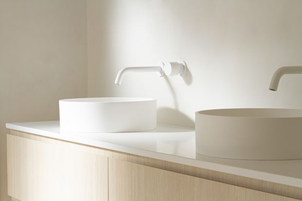 Close-up of a minimalist bathroom vanity featuring two white vessel basins, wall-mounted curved faucets, a pale stone or composite countertop, and light timber-grain cabinetry. Soft natural lighting creates a calm, warm atmosphere.