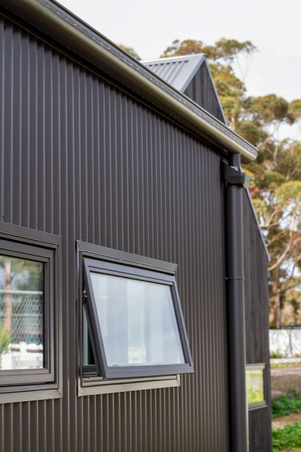 Close-up exterior view of a dark charcoal building clad in vertically ribbed metal panels, with a pitched metal roof, matching gutters and downpipe, and dark-framed windows including one awning window opened outward. Trees and a fenced outdoor setting are visible in the background.
