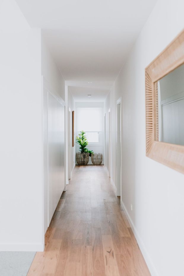 A bright, narrow residential hallway with white walls and ceiling, pale timber flooring, multiple white doorways, and a large timber-framed mirror on the right wall. Potted greenery and woven baskets decorate the far end, creating a clean, airy feel.