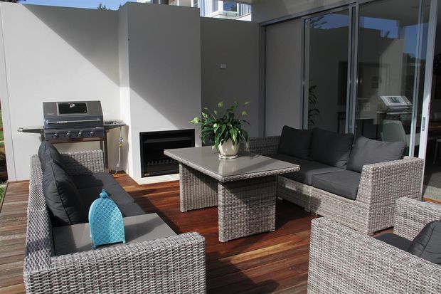A compact, sunlit outdoor entertaining area on a timber deck, furnished with grey woven wicker sofas and armchairs around a central outdoor coffee table. The space includes a built-in barbecue, a freestanding outdoor fireplace, potted greenery, and large glazed sliding doors connecting to the interior.