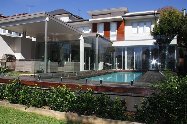 Exterior view of a contemporary multi-level residence with a flat-roofed extension, extensive floor-to-ceiling glazing, timber-clad wall accents, and a raised swimming pool surrounded by a timber deck. A covered outdoor entertaining area and landscaped garden are visible in the foreground.