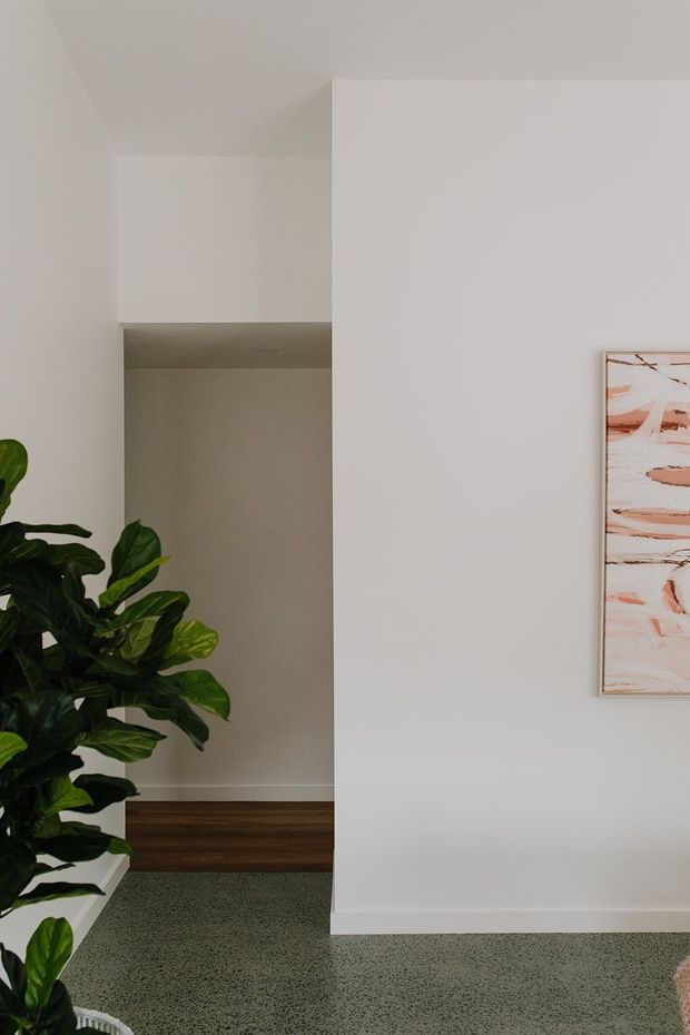 A minimalist interior corner or hallway with white plaster walls, a projecting partition wall, dark timber flooring visible through an opening, and a green terrazzo-like floor in the foreground. A large leafy indoor plant sits at left, while a framed abstract artwork with blush and terracotta marks hangs on the right wall.
