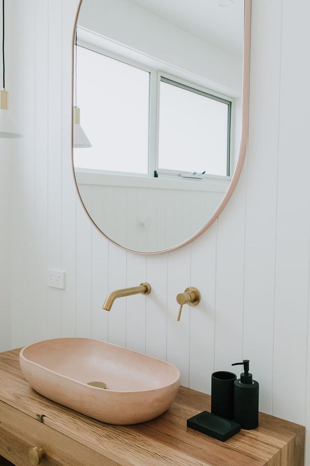 Close-up of a bright, minimalist bathroom vanity featuring a freestanding oval stone basin on a thick timber countertop, wall-mounted brushed brass tapware, black soap dispensers, white vertical wall panelling, and a large rounded mirror. A frosted or bright window provides natural light.