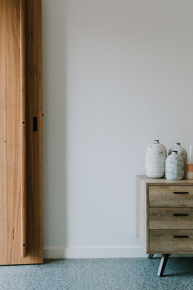 A minimalist interior vignette featuring a full-height natural timber sliding door or wall panel on the left, a pale grey painted wall, and a mid-century-style timber drawer cabinet on tapered dark legs. Textured terrazzo-like flooring extends across the foreground, while ceramic vessels sit on the cabinet.