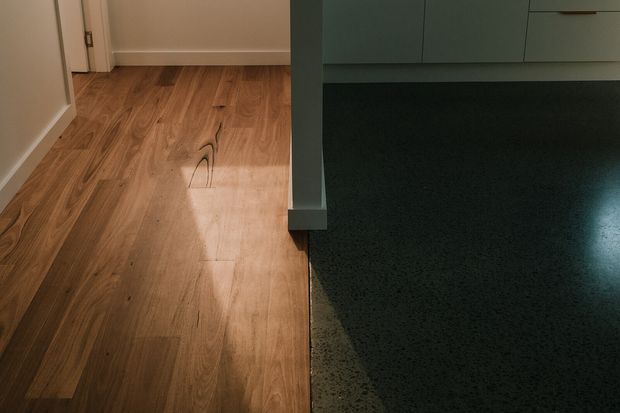 Close-up interior view showing a transition between warm timber flooring in a hallway or adjoining room and a dark polished concrete or terrazzo-like floor beneath pale built-in cabinetry. White painted walls and door trim frame the timber-floored area.