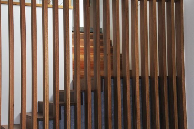 Interior view of a staircase screened by a dense vertical timber batten partition. Dark timber stair treads and risers are visible behind the warm wood slats, with a simple white wall and glimpses of a light-filled landing beyond. The composition highlights the vertical architectural screen and stair structure.