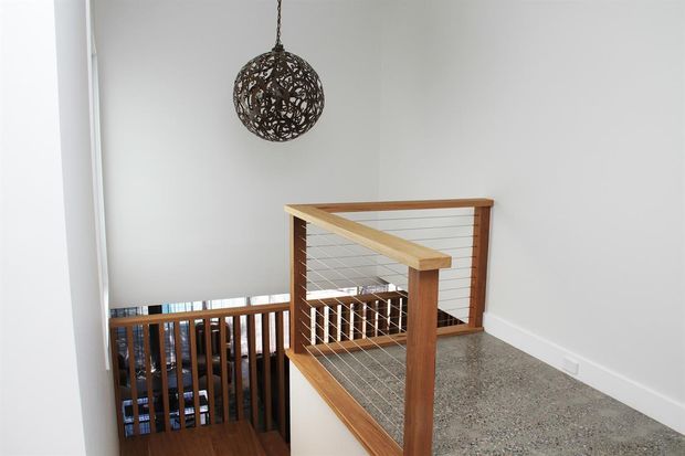 A bright upstairs landing beside an open stairwell, featuring a timber-and-wire balustrade, grey carpeted flooring, white walls, and a dark spherical pendant light. Timber stairs and lower-level glazing are visible through the balustrade.