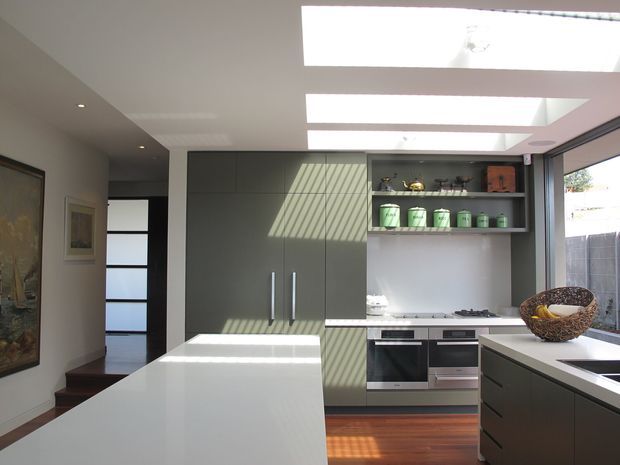 A bright contemporary kitchen with a large white island, dark grey-green full-height cabinetry, integrated stainless-steel appliances, open display shelving, and timber flooring. Three large ceiling skylights provide strong natural light, while a glazed opening and hallway connect to adjacent areas.