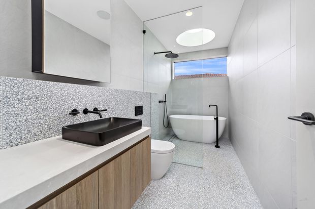 Bright contemporary bathroom with a walk-in shower enclosed by clear glass, freestanding oval bathtub, wall-mounted black fixtures, and a floating timber-veneer vanity with a dark vessel basin. White wall panels, a speckled terrazzo-like floor and splashback, large mirror, skylight, and high window create a clean, light-filled space.