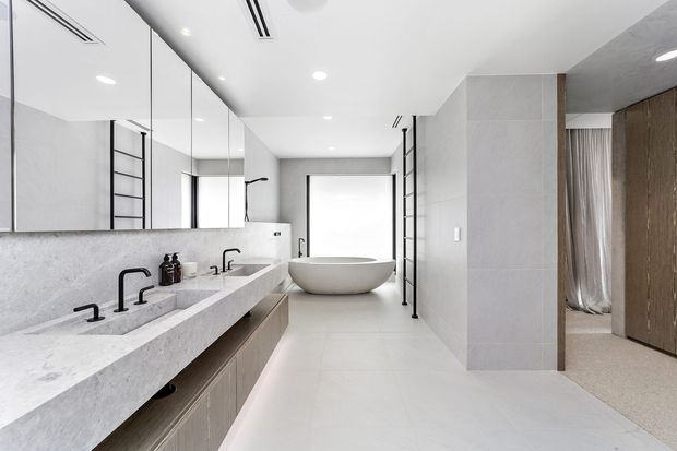 Bright contemporary bathroom with a long double vanity, pale stone benchtop, integrated basins, black tapware, mirrored wall cabinetry, freestanding oval bathtub, large light-toned wall and floor tiles, and black ladder-style towel rails. A carpeted bedroom or dressing area is visible through the opening on the right.