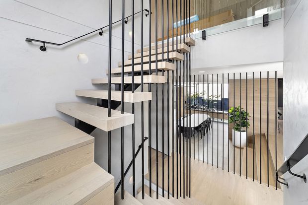 Bright contemporary interior view of a floating stairway with pale timber or timber-look treads, slim black metal balustrade rods, and a matching black handrail. The stair rises through a double-height void beside a mezzanine, with pale timber flooring and wall panelling, white walls, and glimpses of a dining area and greenery beyond. The open vertical balustrade creates a strong linear architectural feature.