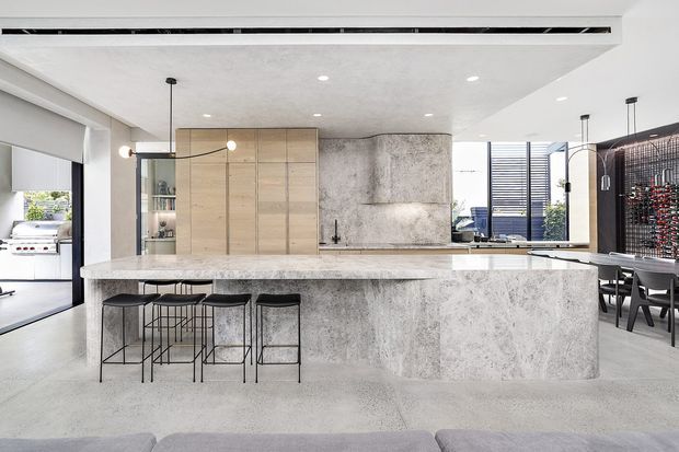 A bright, open-plan contemporary kitchen with a large monolithic stone island and matching stone splashback. The island includes a long integrated benchtop with black metal bar stools, while pale timber cabinetry provides concealed storage. Black-framed glazing, pendant lights, recessed ceiling lighting, and a dining area extend the space toward the rear. An outdoor barbecue area is visible through the glazing on the left.