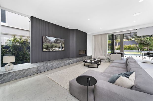 Contemporary open-plan living room with a large curved grey sectional sofa, pale carpet, and a dramatic dark vertical-panelled feature wall incorporating a television and linear fireplace. A long grey stone hearth extends beneath the wall, while full-height glazing and sliding doors open toward a landscaped outdoor area. Neutral furnishings, recessed lighting, and black-framed windows create a refined, spacious interior.
