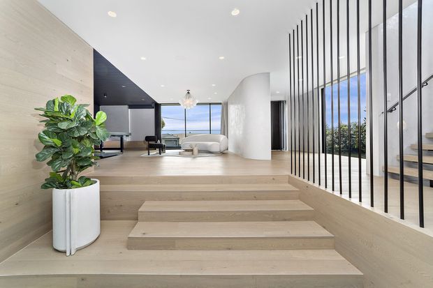 Bright contemporary open-plan interior featuring broad pale timber steps leading to a raised living area. The space has light timber wall and floor finishes, white curved architectural elements, recessed ceiling lighting, floor-to-ceiling glazing, and a black vertical metal screen separating the stair and living zones. A white sofa and indoor plant add furnishings to the refined, airy space.