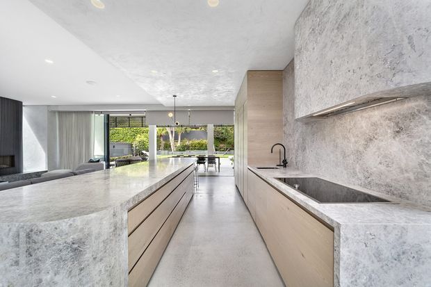 A bright, open-plan contemporary kitchen with a long central island and matching perimeter cabinetry in pale timber tones. The kitchen features extensive grey-white stone surfaces, including the island, benchtops, splashback and integrated sink area, with a black tap and understated pendant lighting. Floor-to-ceiling glazing at the rear opens the space toward a landscaped outdoor area, while a living area is visible to the left.