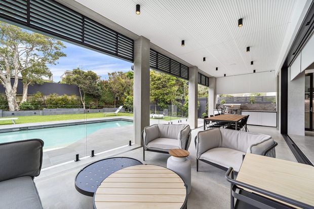A bright, covered outdoor entertaining area beside a swimming pool, furnished with contemporary lounge chairs, a round coffee table and a dining table. The space features a white slatted ceiling with recessed lighting, concrete structural columns, dark louvred screening, large glazed openings with frameless pool fencing, and views to landscaped grounds.