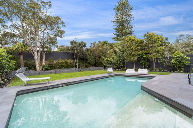 A bright landscaped backyard with a large modern swimming pool, shallow entry area, pale grey tiled and paved surrounds, sun loungers, manicured lawn, mature trees, and dark boundary fencing. The scene has a clean contemporary outdoor entertaining feel under a clear blue sky.