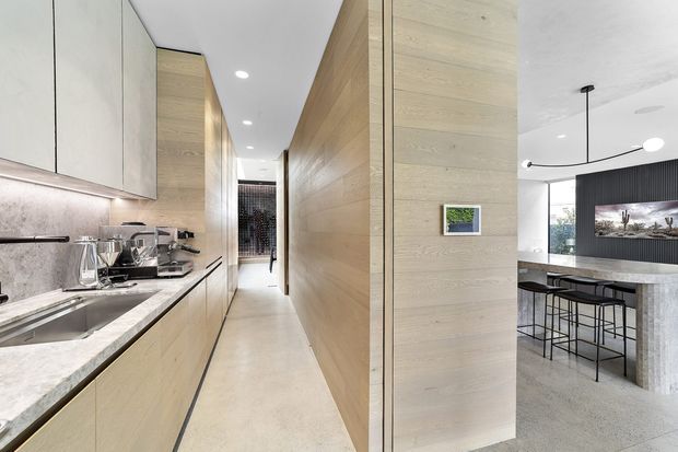 A contemporary open-plan kitchen and circulation area with pale timber cabinetry and wall panelling. The galley kitchen has a light stone benchtop, integrated sink, appliances and under-cabinet lighting, leading toward a hallway. Beyond the timber-lined partition is a refined dining or bar area with a stone island, black stools, dark ribbed feature wall and minimalist pendant lighting.