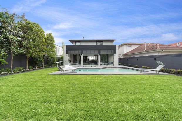 Wide view of a contemporary flat-roofed home from a landscaped backyard, with a dark-and-white rendered facade, extensive rear glazing, a rectangular swimming pool, paved pool terrace, boundary walls, and a large manicured lawn.