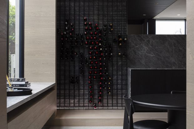 A dark, refined contemporary dining and wine display area. A floor-to-ceiling black metal wine rack forms a feature wall, with bottles arranged throughout. The space includes a round black dining table and chairs, dark stone benchtop and splashback surfaces, light timber cabinetry and wall panels, and recessed ceiling lighting.