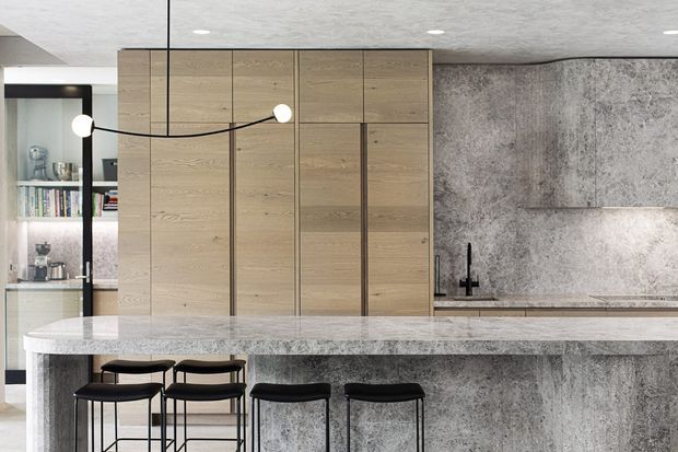 Contemporary kitchen with a long stone island and integrated breakfast bar, black stools, tall light timber cabinetry, and a matching textured grey stone splashback and island base. A black-framed glazed display cabinet and minimalist pendant lighting add contrast.