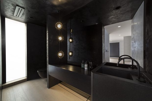 A dramatic, dark-toned contemporary bathroom with a large black vanity and integrated basin, sculptural black tapware, dark textured wall and ceiling finishes, pendant globe lights, and pale timber flooring. A toilet is partially visible beside a bright frosted window, while an open doorway leads to a lighter adjacent space.