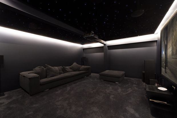 A dark, sophisticated home cinema with a large sectional sofa and ottoman, deep charcoal carpeting and walls, concealed perimeter LED lighting, a star-like fibre-optic ceiling, and a projector mounted overhead. A large screen or display wall is visible at the front, with media equipment along the side.