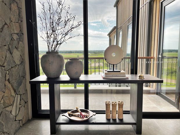 A styled console table sits in a modern interior beside full-height glazing, with sculptural ceramic vessels, books, a round decorative object, and small accessories. A rough natural-stone wall contrasts with dark metal-framed windows and doors, while a broad rural landscape is visible outside.