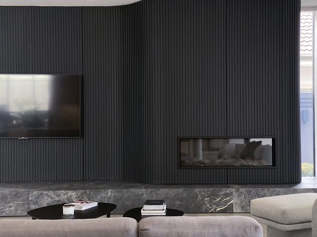 Contemporary living room feature wall with dark vertical fluted panelling, an inset linear fireplace, wall-mounted television, and a dark marble hearth or ledge. Neutral upholstered sofas and low black coffee tables sit in the foreground, with daylight entering from the side.