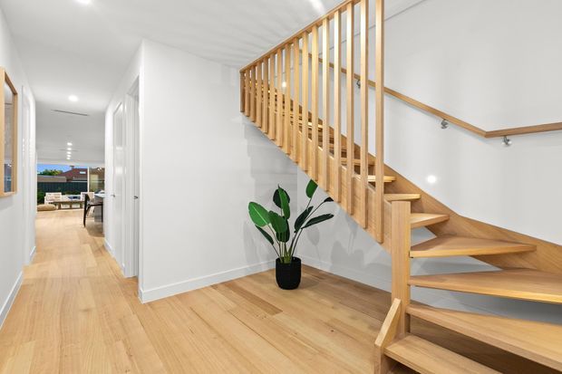 Bright contemporary residential hallway with a timber staircase, open vertical balustrade, white walls, recessed ceiling lights, and light timber flooring. A corridor leads toward a living area at the rear, with a potted plant beneath the stairs.