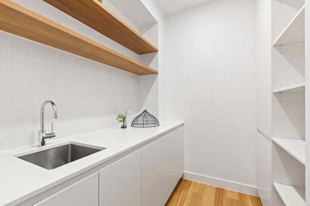 Bright, compact contemporary kitchen or butler’s pantry with white cabinetry, a white benchtop, stainless-steel undermount sink and chrome tapware. Open light timber shelving runs above a vertically textured white splashback, with additional white open shelving on the right. Warm timber flooring contrasts with the predominantly white fit-out.