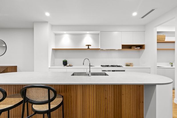 Bright contemporary kitchen viewed from behind a large island. The space features white full-height cabinetry, integrated shelving with timber accents, a pale stone benchtop, a timber-lined island with a rounded edge, stainless steel sink and tapware, and recessed ceiling lighting. Cane and black-frame stools sit at the island, with a pantry or shelving area visible at right.