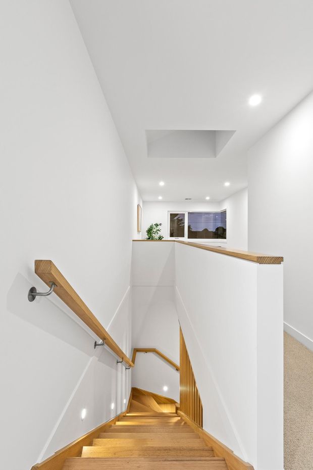 Bright minimalist stairwell viewed from an upper landing, with a long timber staircase descending through a narrow white-walled void. Timber handrails, open balustrade detailing, recessed ceiling lights, a skylight or ceiling opening, and a small window at the lower landing create a clean, contemporary interior.