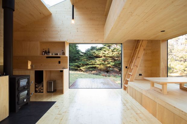 A compact timber-lined cabin interior with a wood-burning stove, built-in open shelving and cabinetry, a skylight, and a large glazed opening onto a timber deck and wooded landscape. A raised built-in seating or sleeping platform with ladder occupies the right side, while a simple built-in table and bench extend into the room. The space has a warm, minimalist, nature-oriented character.
