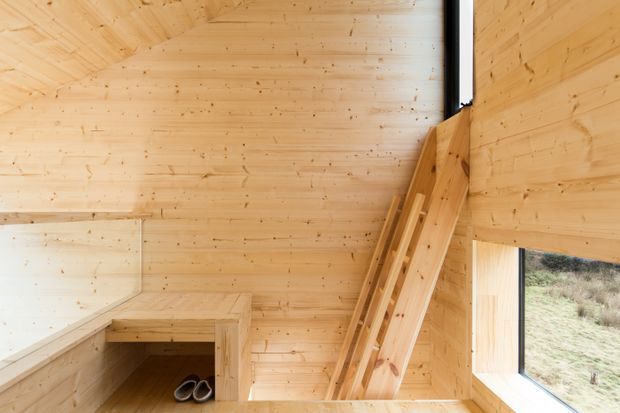 Compact timber-lined interior with pale wood wall and ceiling panelling, a built-in bench or storage unit, and a steep timber ladder leading upward. Large corner glazing provides views to the natural landscape outside. The space has a simple cabin-like, minimalist character and appears to function as a small entry or circulation area.