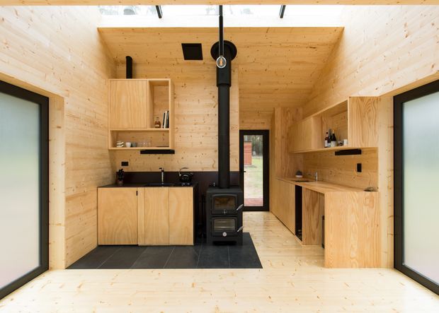 Compact timber-lined interior with a kitchenette and built-in wooden cabinetry on both sides, a black tiled cooking area, and a freestanding black wood-burning stove with flue centered in the room. Pale timber walls, ceiling and floor create a warm minimalist cabin-like space. Large frosted-glass sliding doors flank the room, with a glazed door at the rear.