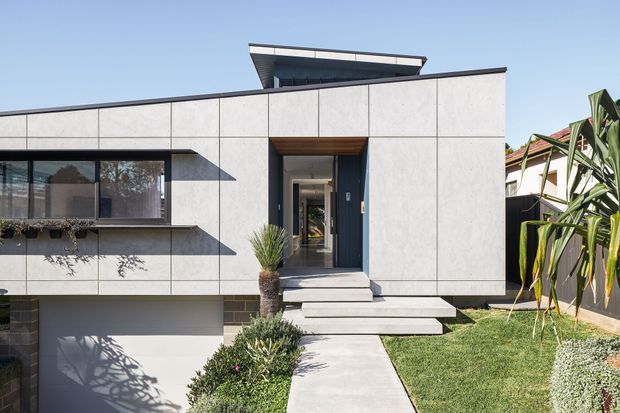 Front exterior view of a contemporary single-storey residence with a flat roof, pale panelled facade, dark-framed glazing, recessed entry, timber-lined porch soffit, and stepped concrete path. Low-maintenance landscaping and a partially visible garage complement the clean geometric design.