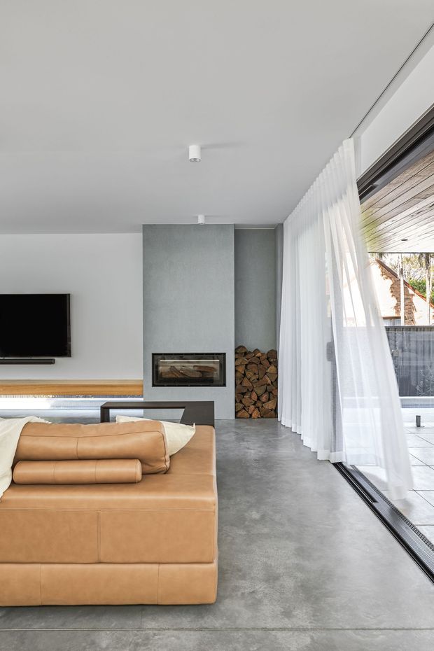 A bright, minimalist living room with a large tan leather sectional sofa, polished grey concrete flooring, white walls and ceiling, and expansive floor-to-ceiling glazing screened by sheer white curtains. A dark grey central feature wall contains a built-in fireplace and neatly stacked firewood, while a timber-lined covered outdoor area is visible beyond the glazing.