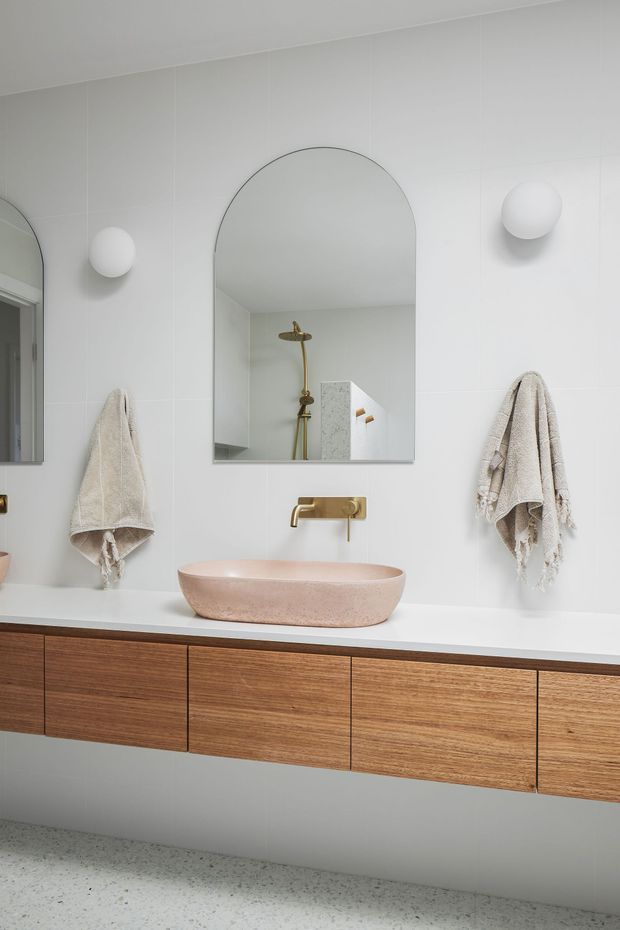 A bright, refined contemporary bathroom vanity featuring a wall-mounted mid-wood timber vanity with flat-front drawers, a white benchtop, and a pale blush-pink vessel basin. White tiled walls, an arched mirror, rounded wall sconces, and brushed brass tapware create a clean, softly curved composition. A shower with brass fittings is visible in the mirror reflection, while textured neutral towels hang on either side.
